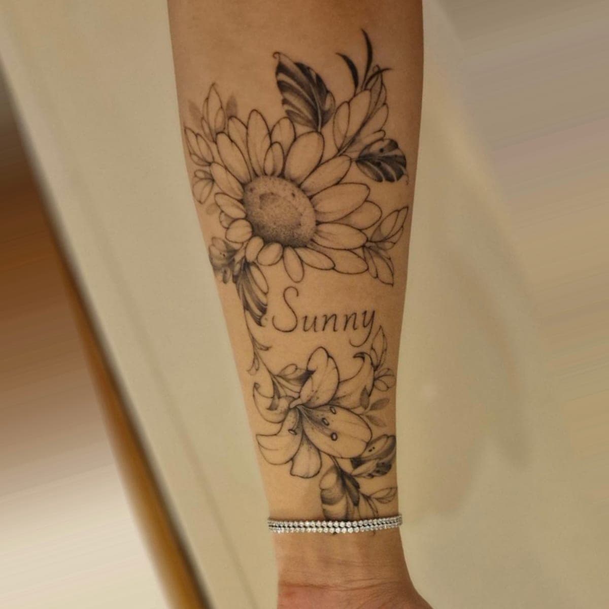 Healed black and grey sunflower and lilies with the name Sunny on a forearm