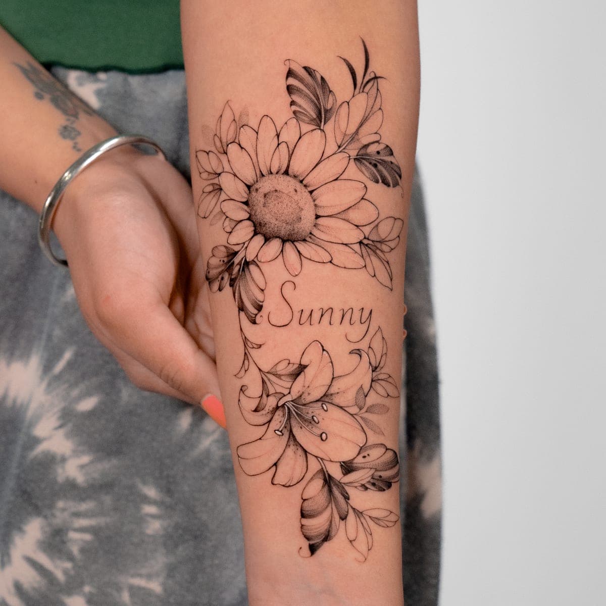 Fresh black and grey sunflower and lilies with the name Sunny on a forearm