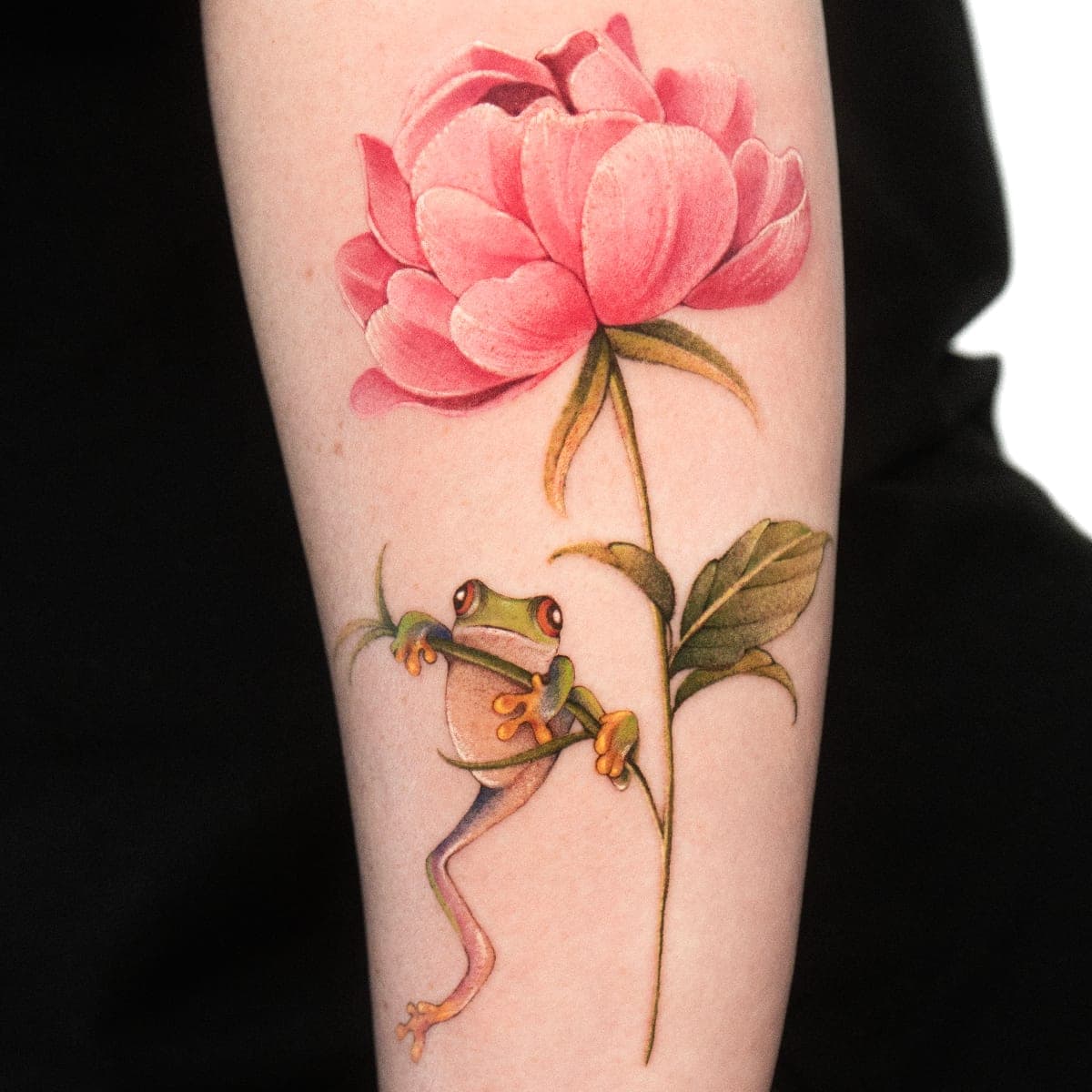 Fresh colour tattoo of a pink peony with a tree frog climbing its stem on a forearm