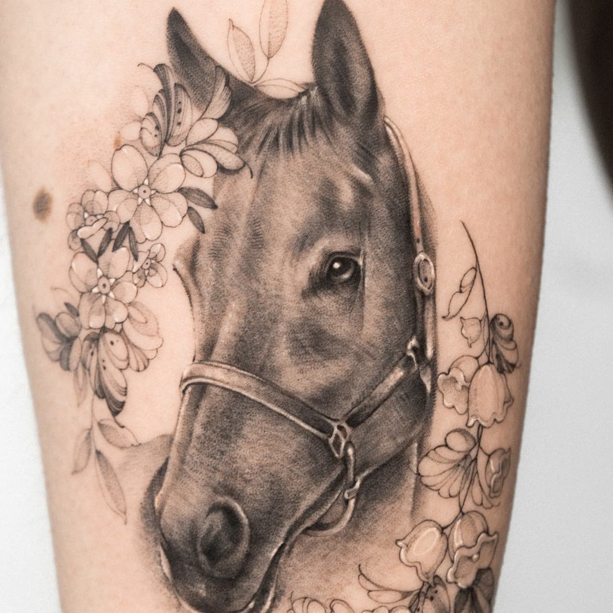 Black and grey tattoo of Boo in her headcollar, framed by flowers, on a forearm